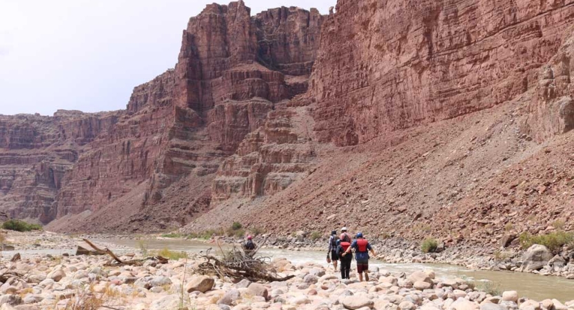 A few people walk a rocky path alongside a river with tall, red canyon walls on the opposite side. 